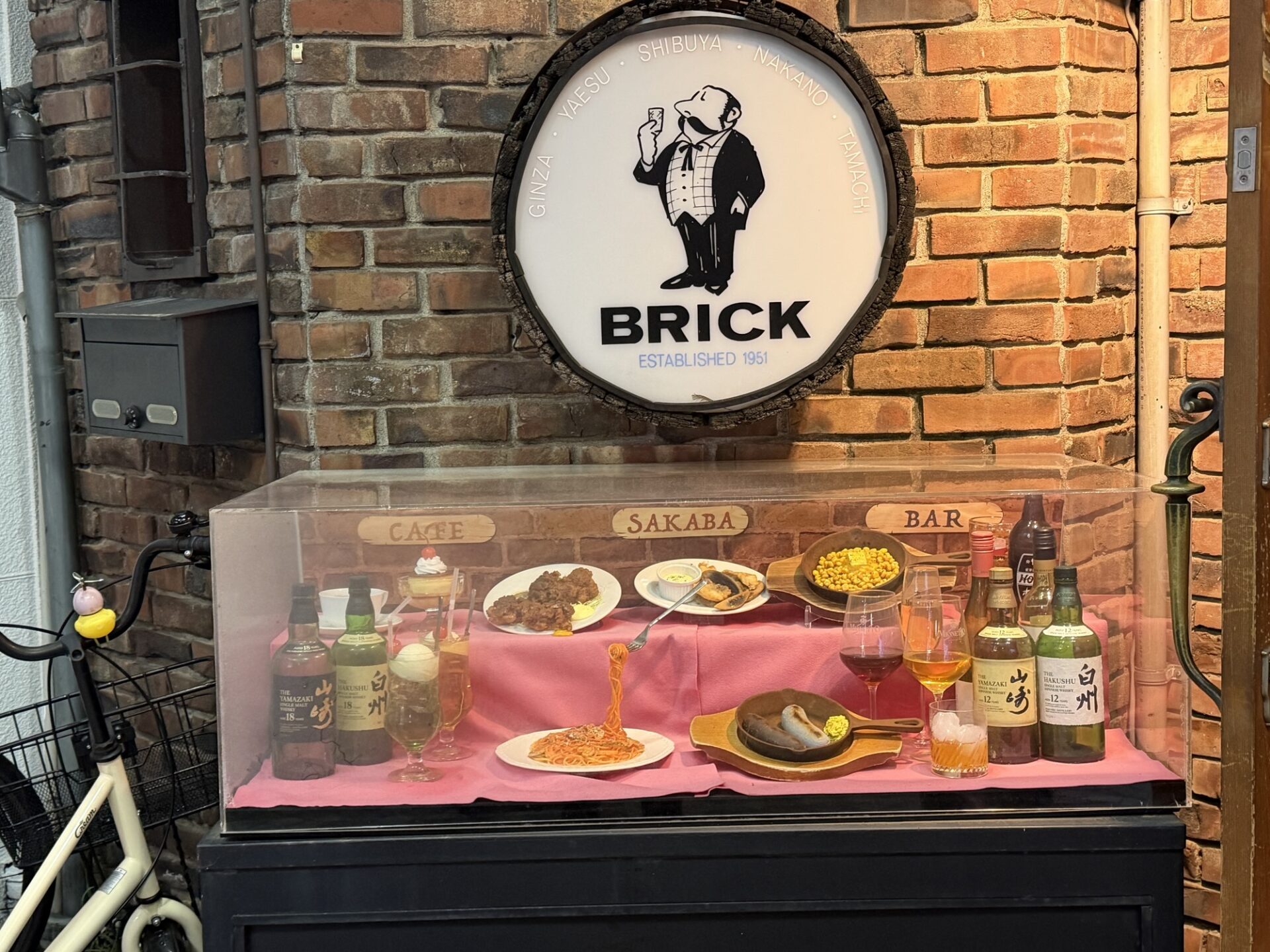 Nakano Brick's round bartender logo on the brick wall above a glass showcase of food and drink samples at the entrance