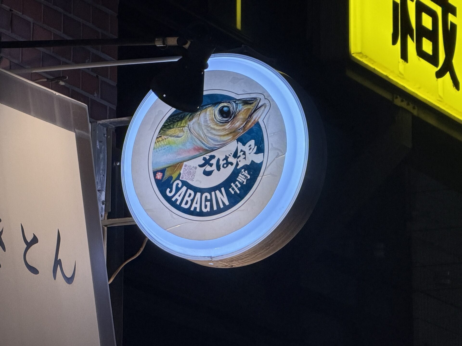 Saba-Gin's round backlit sign at night, featuring a mackerel illustration above the name in Japanese and English